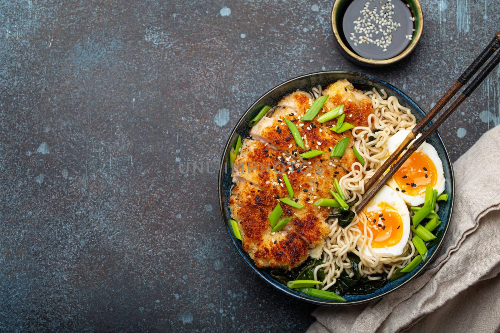 Asian noodles ramen soup, deep fried panko chicken fillet and boiled eggs in ceramic bowl with chop sticks and soy sauce on stone rustic background top view, space for text by its_al_dente