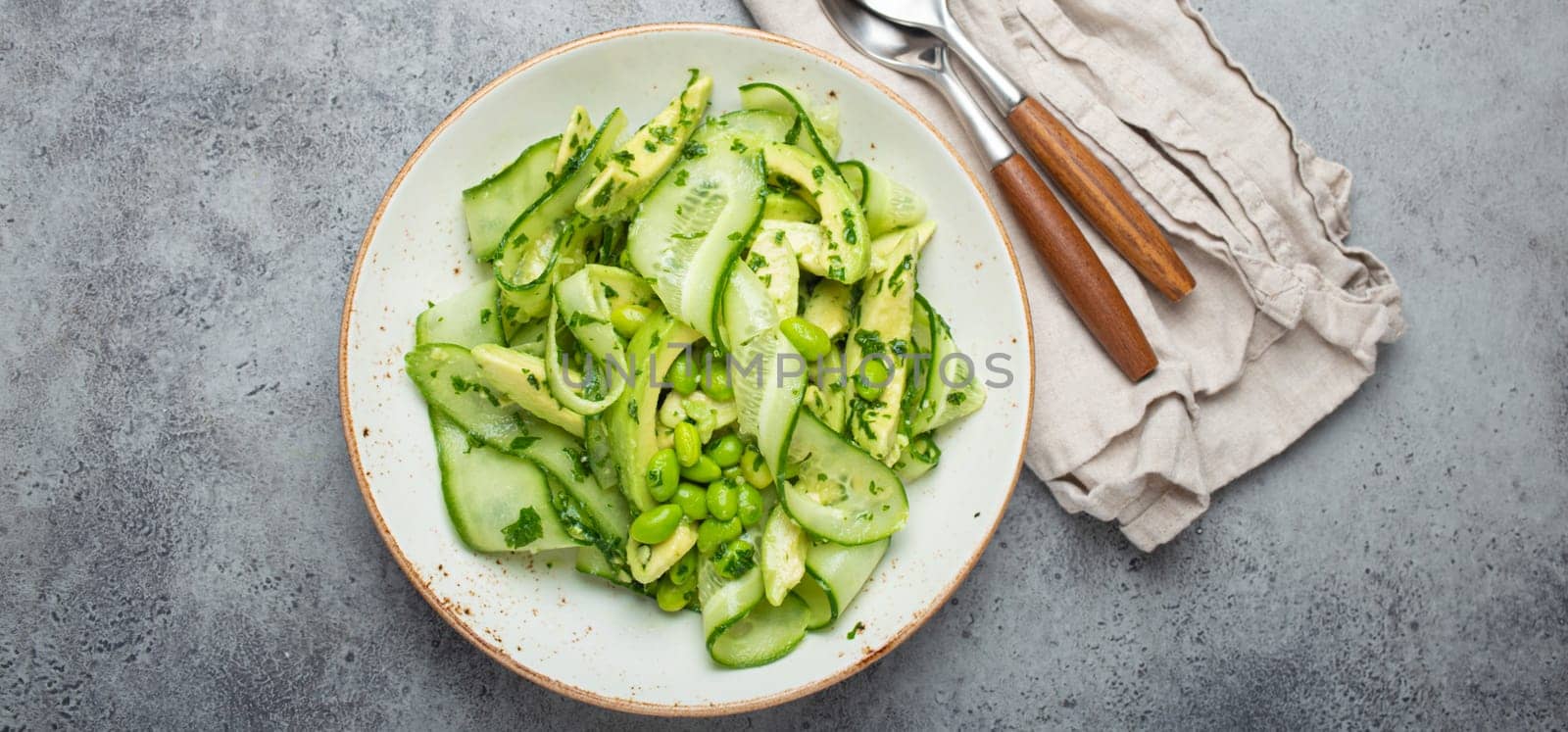 Healthy vegan green avocado salad bowl with sliced cucumbers, edamame beans, olive oil and herbs on ceramic plate top view on grey stone rustic table background