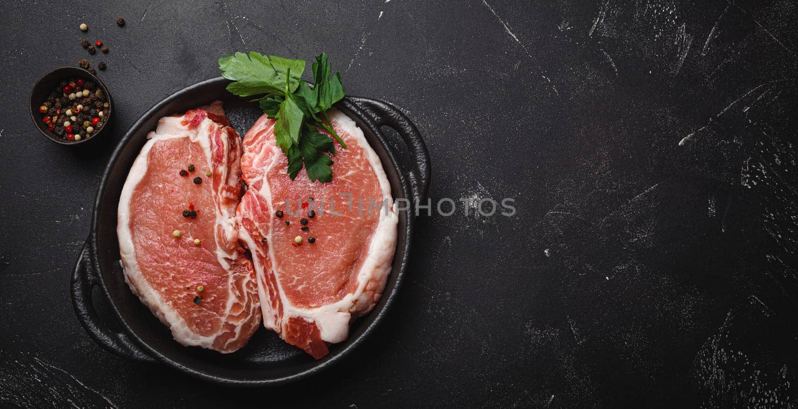 Cut raw meat pork steaks with seasonings in black cast iron pan, dark rustic stone background top view, ready for roasting. Pork loin chops cooking, space for text by its_al_dente