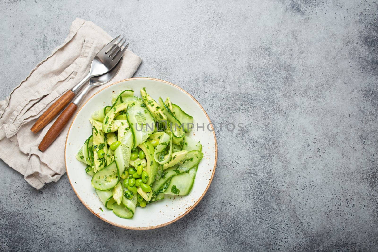 Healthy vegan green avocado salad bowl with sliced cucumbers, edamame beans, olive oil and herbs on ceramic plate top view on grey stone rustic table background. Space for text