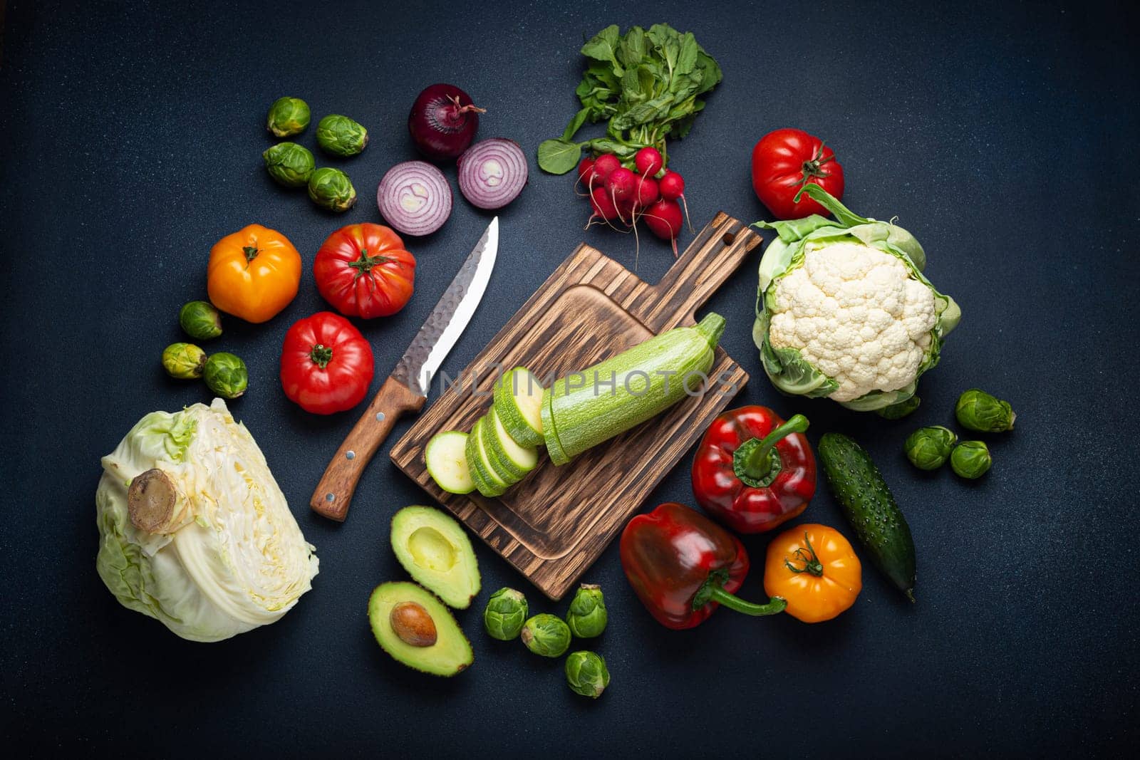 Fresh various vegetables, cut zucchini on wooden cutting board and knife on rustic dark background top view. Cooking vegetarian meal with healthy ingredients, diet food and nutrition concept by its_al_dente