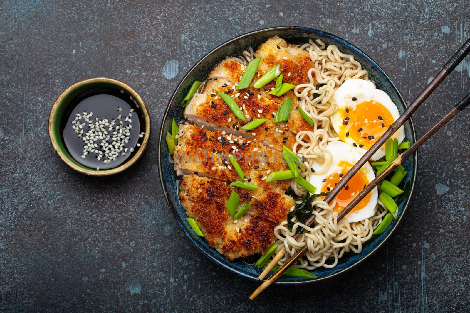 Asian noodles ramen soup with deep fried panko chicken fillet and boiled eggs in ceramic bowl with chop sticks and soy sauce on stone rustic background top view