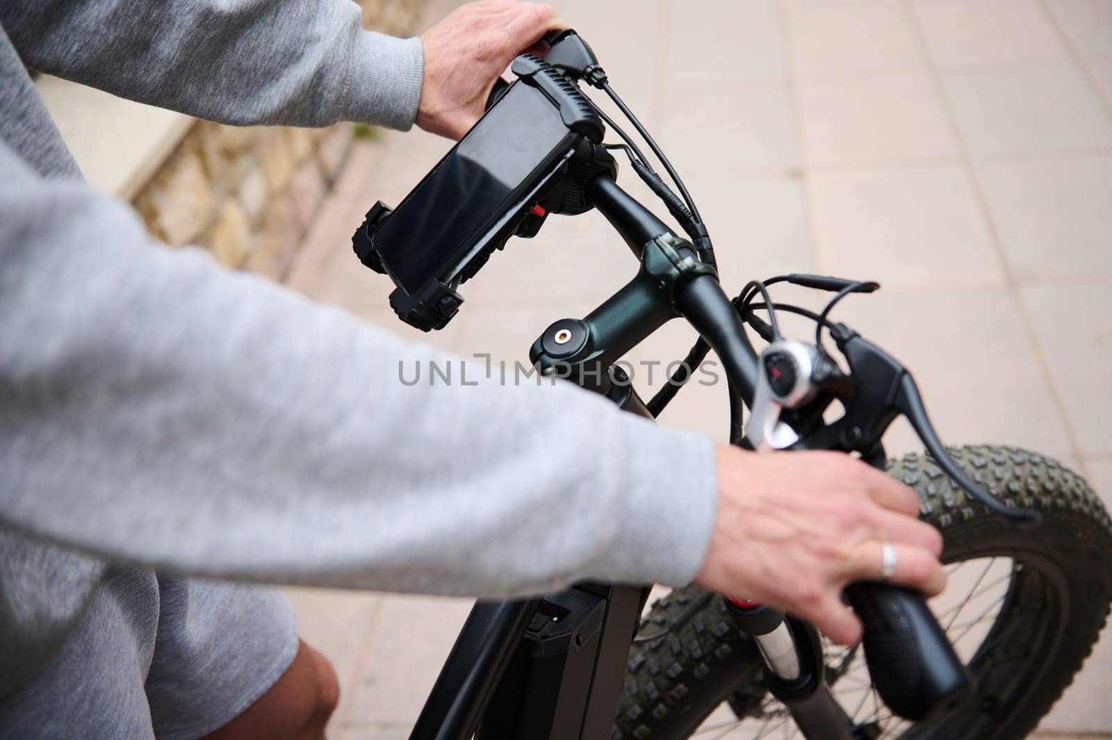 Hands on handlebar of electric bike while riding in city. Smartphone with black mockup screen copy space. Man using mobile phone, renting electric bike using rental app. Bike sharing city service