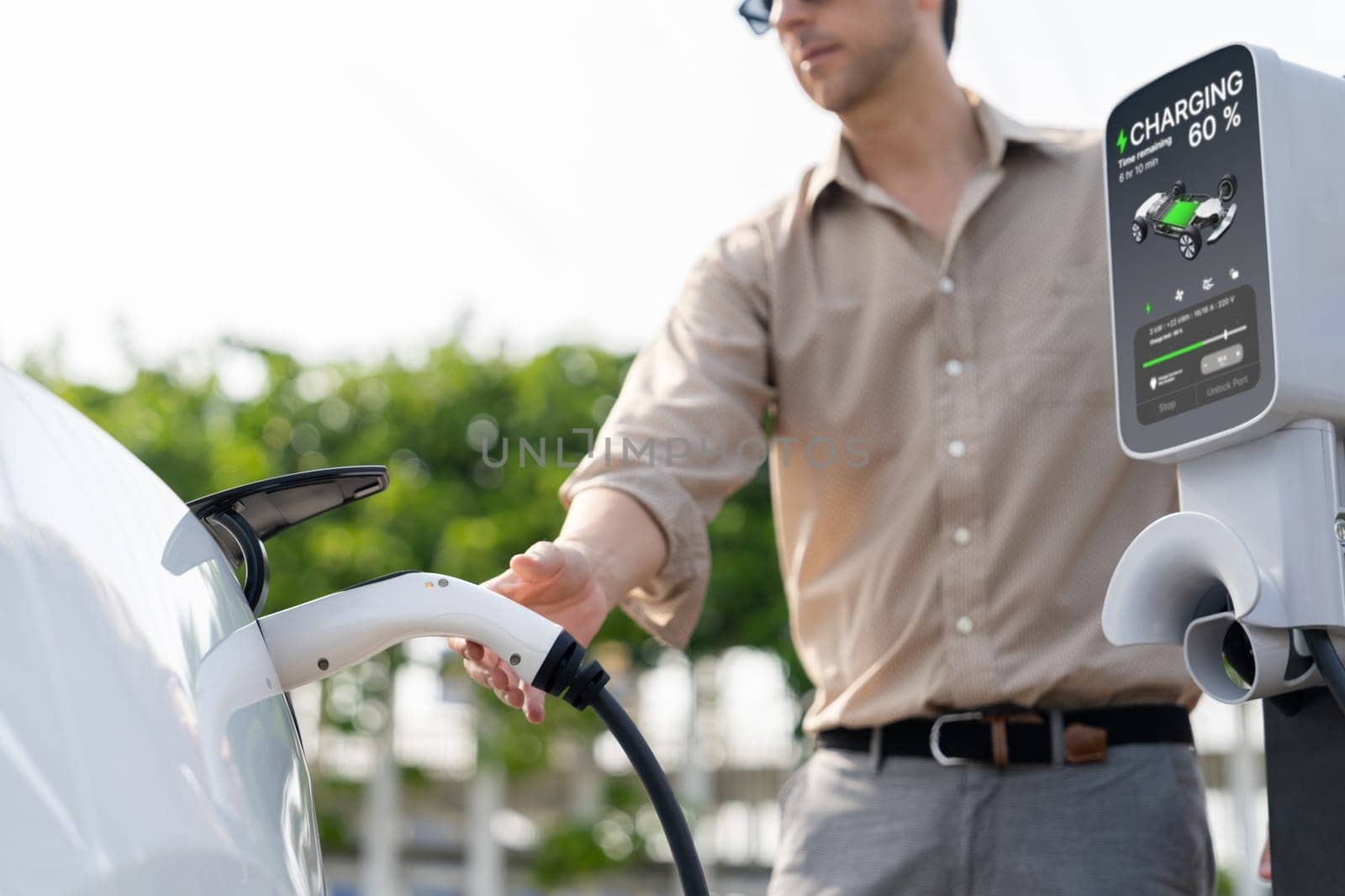 Young man put EV charger to recharge electric car's battery from charging station in city commercial parking lot. Rechargeable EV car for sustainable environmental friendly urban travel. Expedient