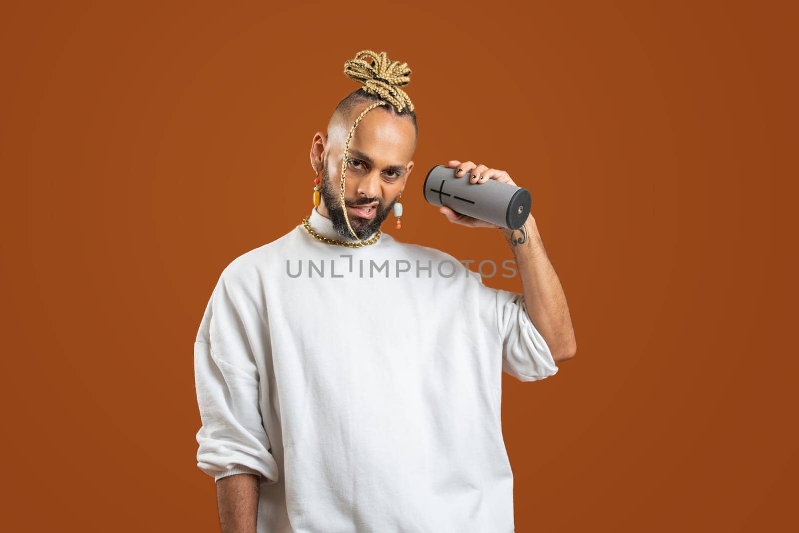 Confident black gay man with bright makeup listen musiv hold in hand portable wireless sound speaker standing isolated on orange background, dressed white. Exudes sense of pride and individuality.
