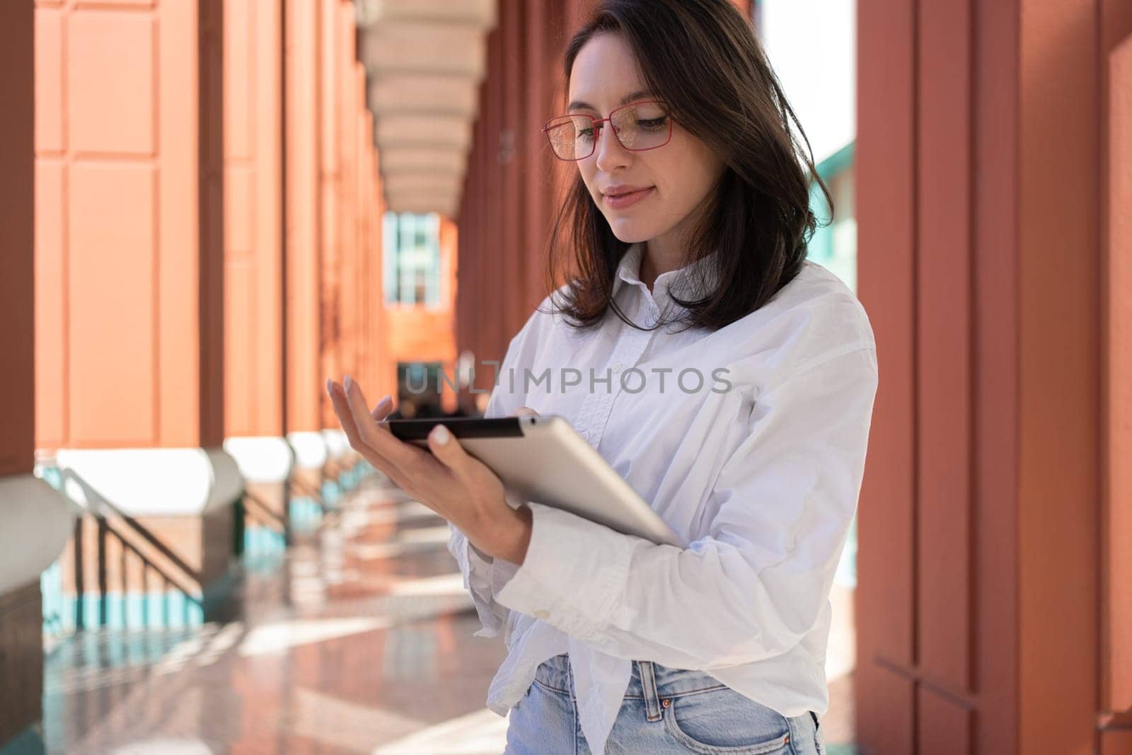 Businesswoman in glasses using digital tablet touching screen, standing outdoor dressed white shirt. Female freelancer holding tablet while walk outside