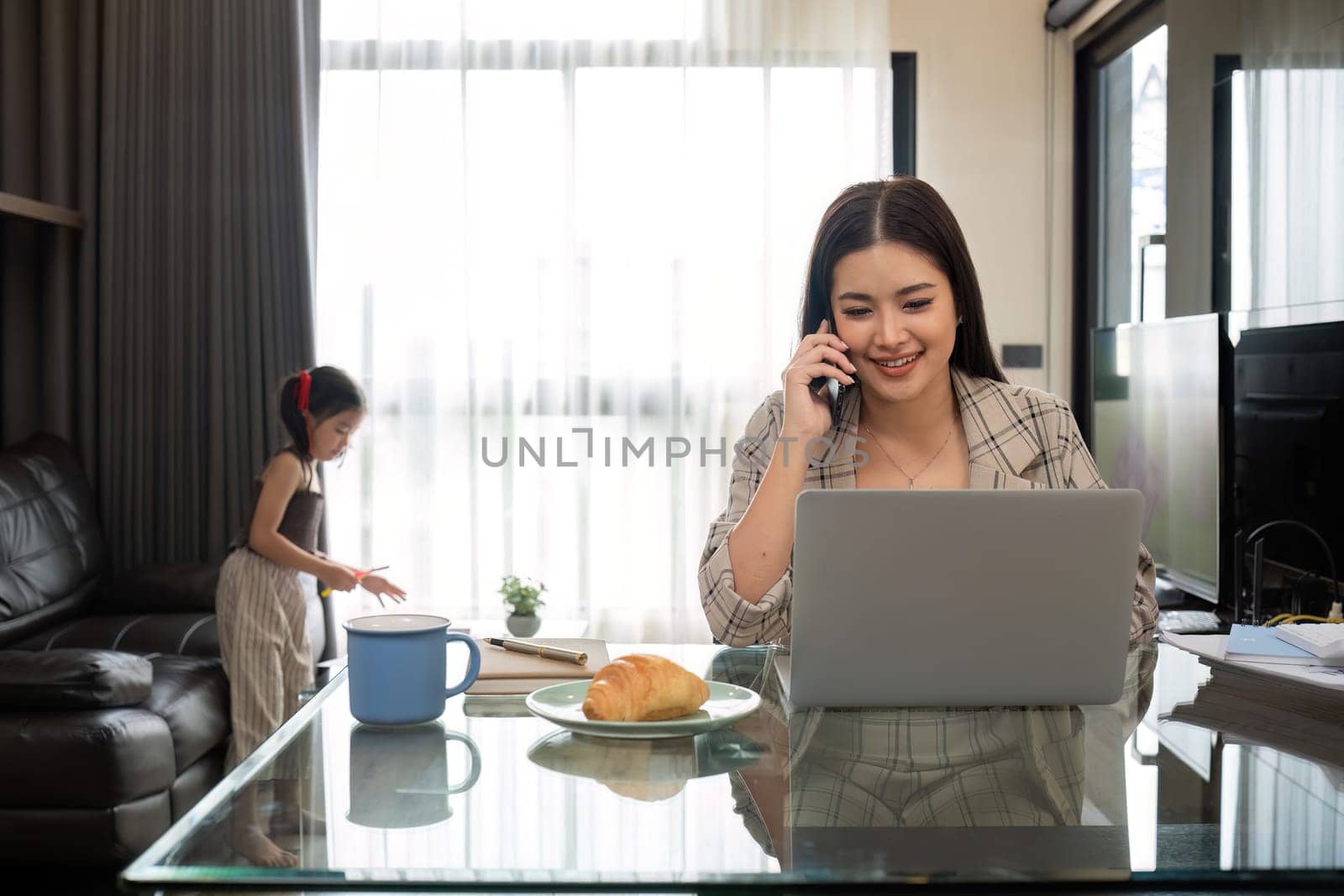 The young woman works from home, raises her daughter, and discusses work with co-workers on the phone and online. by wichayada