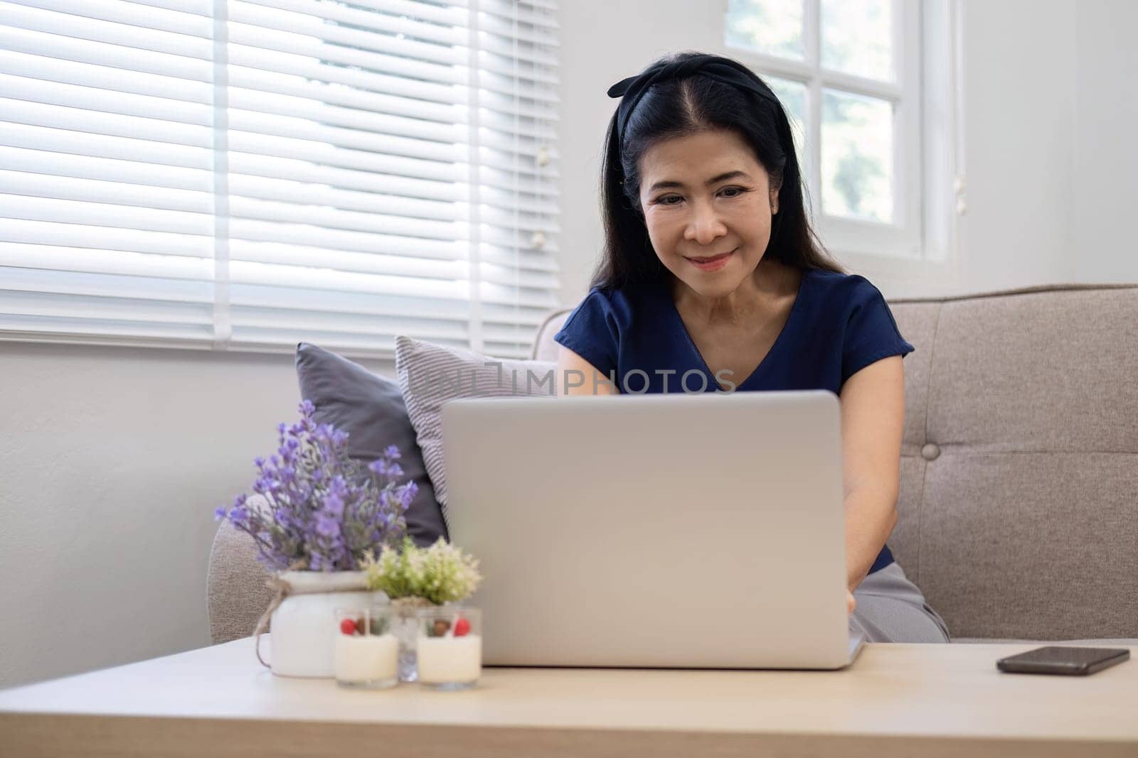 Happy elderly Asian woman in her 60s enjoys using social media on her laptop while relaxing in the living room..