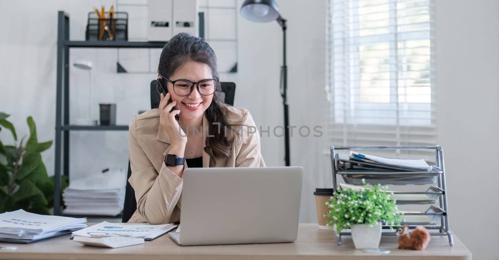 Young Asian business woman sits on the phone in an online business meeting using a laptop in a modern home office decorated with shady green plants..