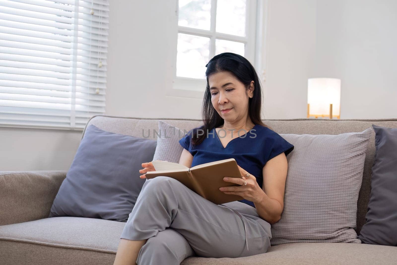 An elderly woman's leisure day sitting and reading a book, relaxing on the sofa in the living room..