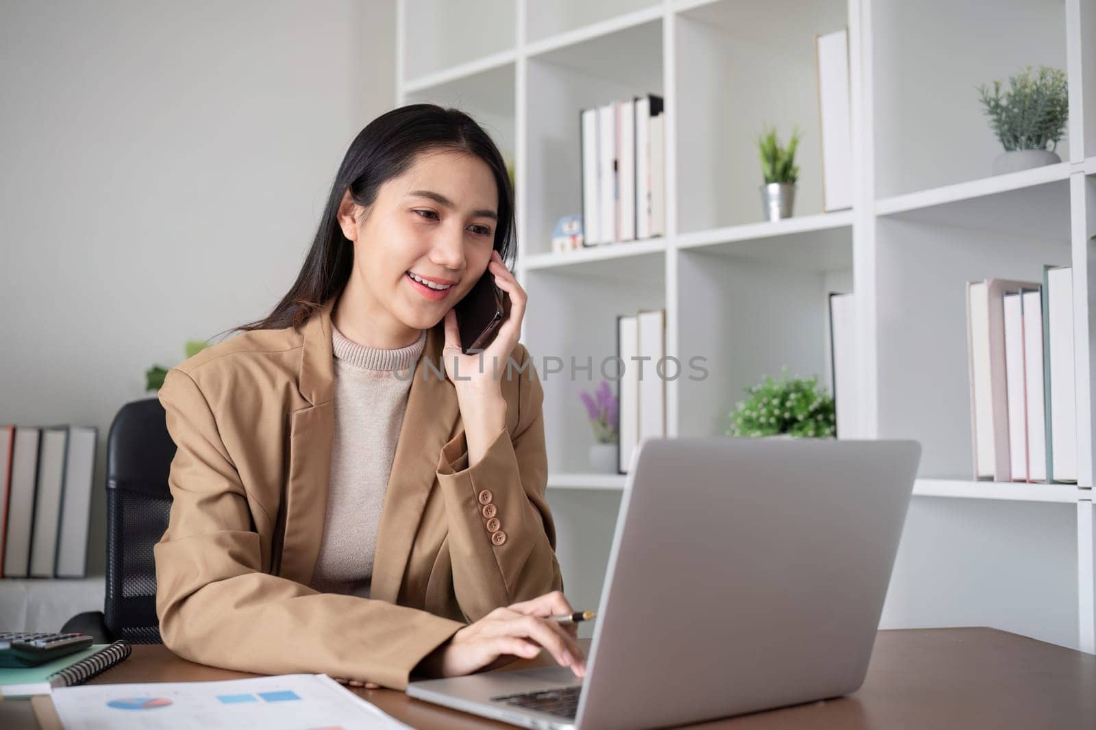 Young Asian business woman sits on the phone in an online business meeting using a laptop in a modern home office decorated with shady green plants..