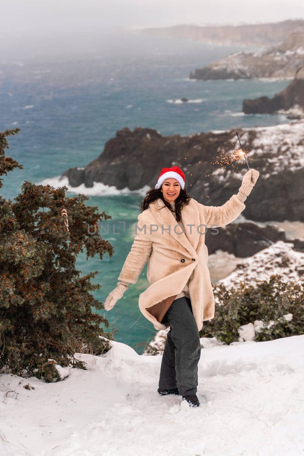 Outdoor winter portrait of happy smiling woman, light faux fur coat holding heart sparkler, posing against sea and snow background.