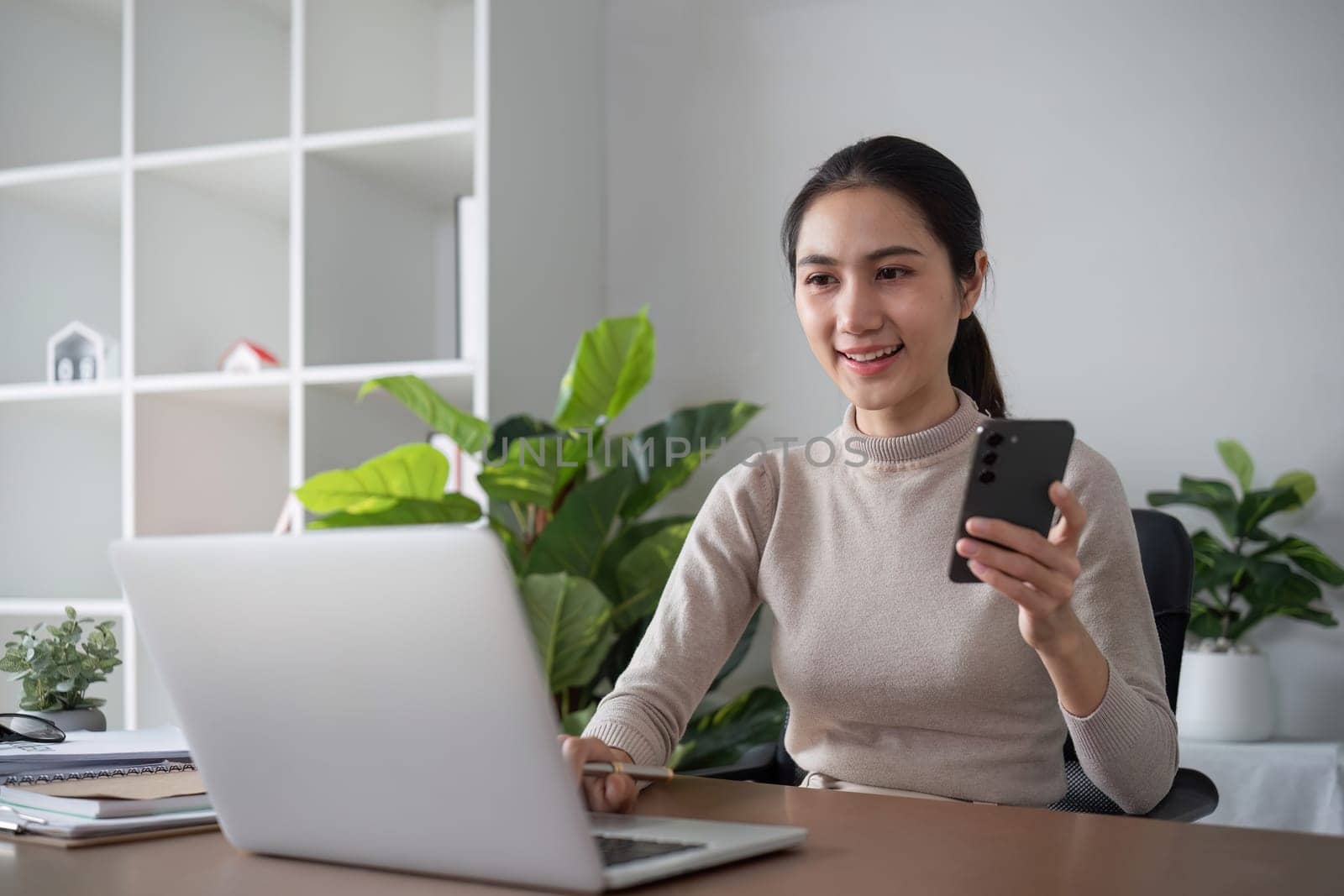 Asian businesswoman or accountant happily sitting and working with laptop on finance and business administration. On the desk in the office decorated with green plants. by wichayada