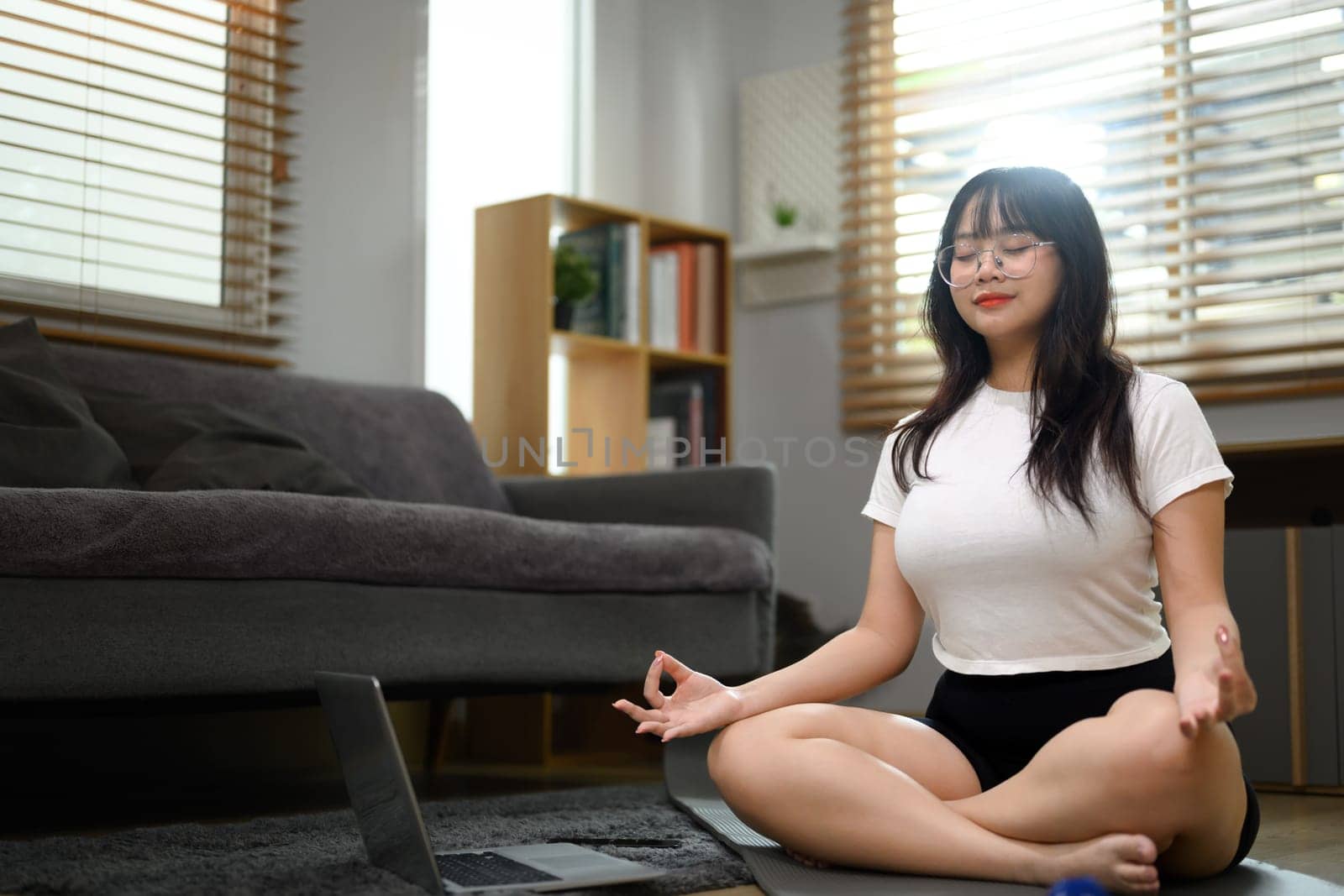 Peaceful young woman meditating in yoga lotus pose with closed eyes on mat at home by prathanchorruangsak