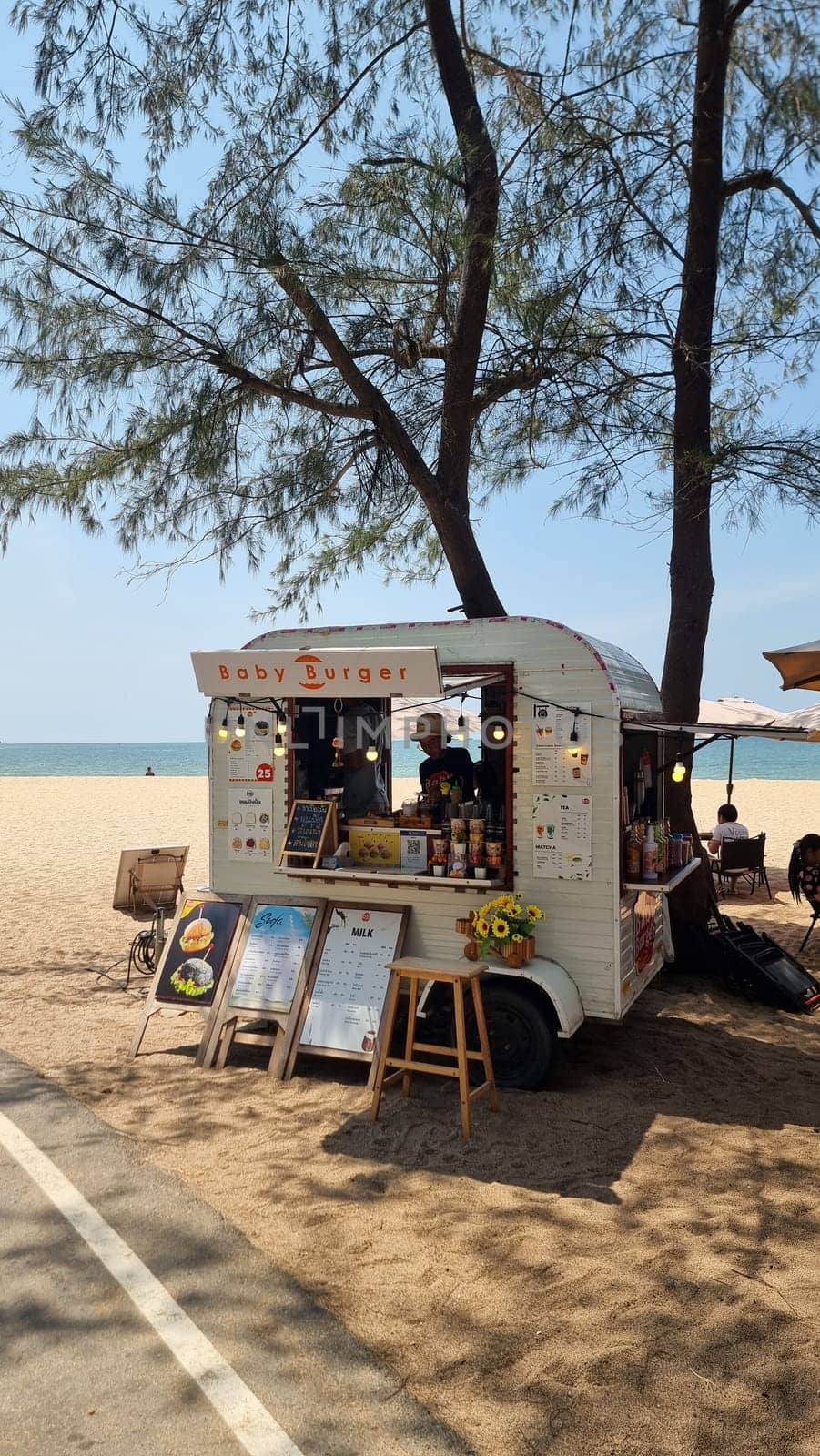 A vibrant food truck is parked on a sandy beach next to a lush tree, serving up delicious treats to beachgoers under the clear blue sky by fokkebok