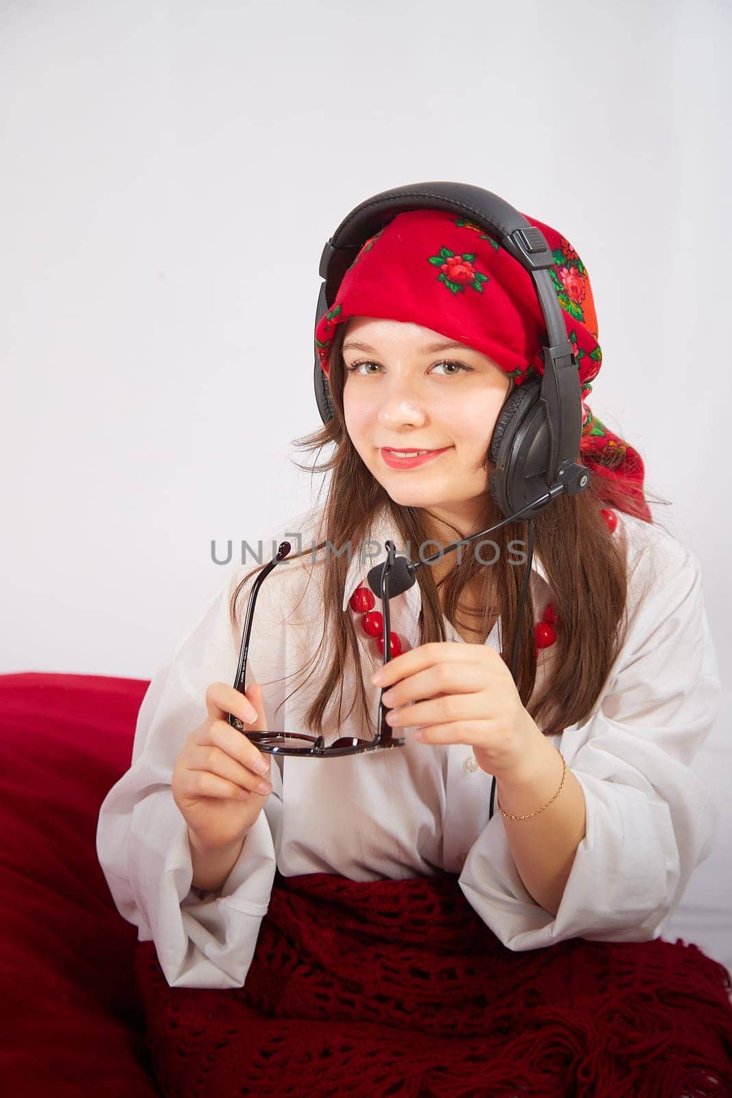 Portrait of young girl in red scarf, large headphones with microphone and black glasses. Woman who is radio or television presenter in workplace. Funny female telecom operator. Freelancer at work