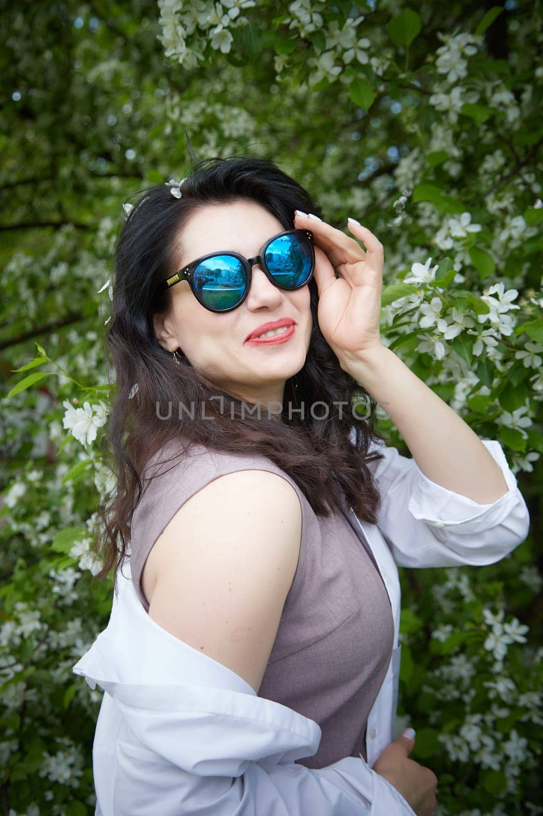 Funny brunette woman in sunglasses near Blossoms of apple tree in Spring Garden outdoors. Concept of face and body care. Scent of perfume and tenderness by keleny