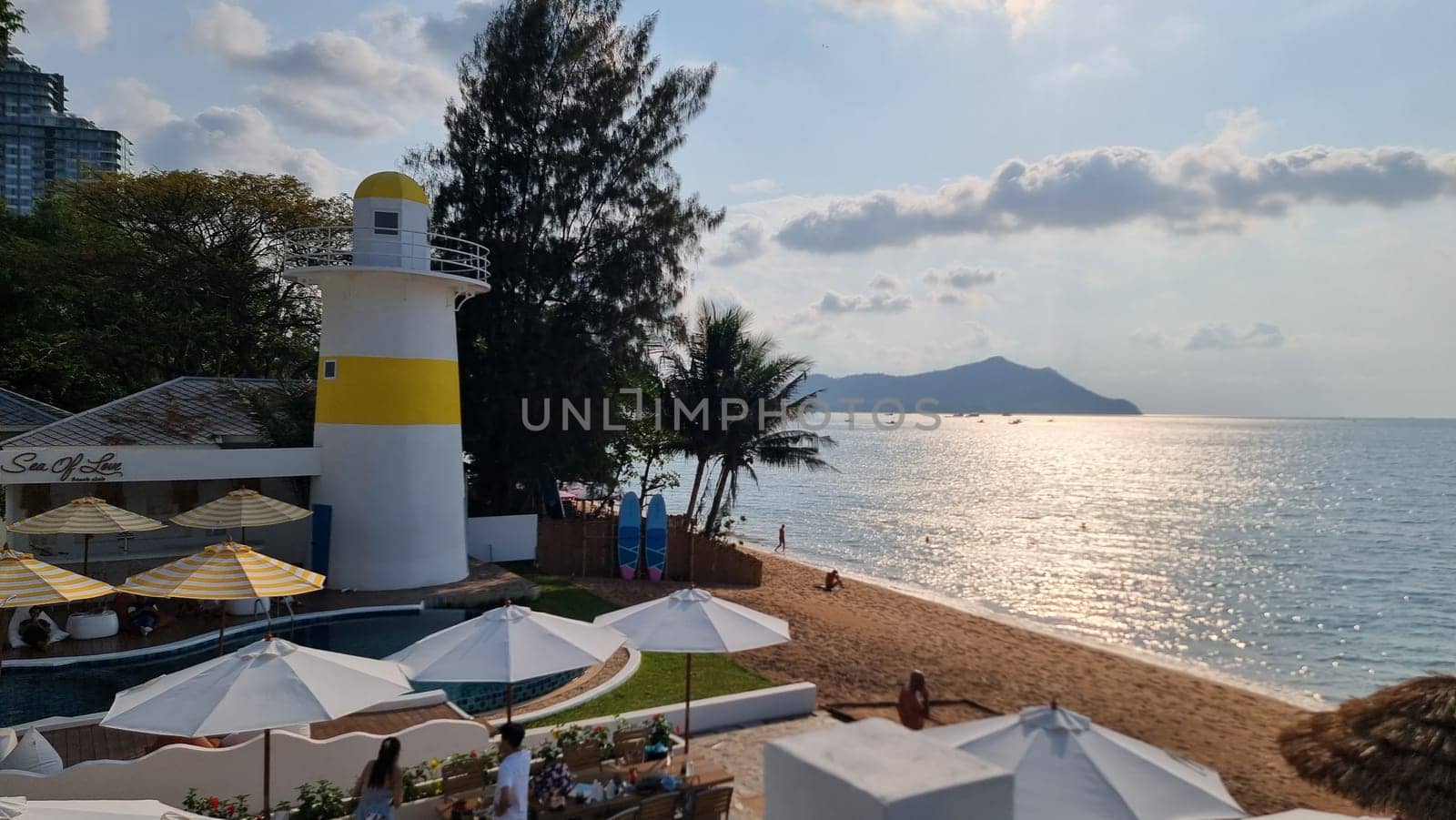 A picturesque scene of a vibrant beach with colorful umbrellas scattered along the shoreline, leading the eye towards a majestic lighthouse standing tall in the distance by fokkebok