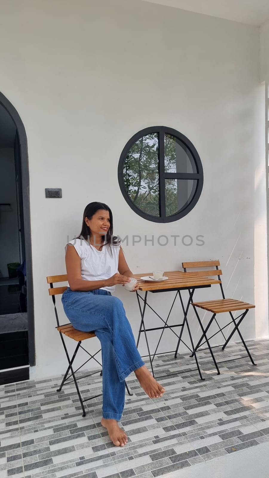 A woman sits peacefully at a table outside a charming house, surrounded by nature, lost in thought.