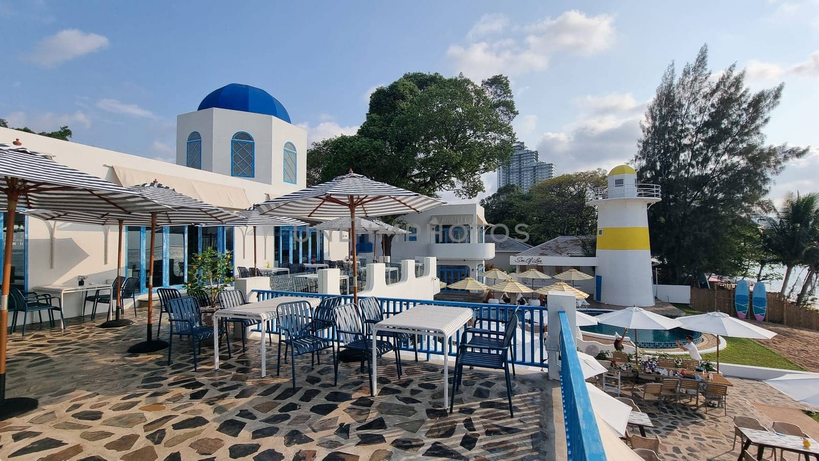 Relaxing outdoor dining area with shaded tables and colorful umbrellas set against a backdrop of lush greenery and clear blue skies by fokkebok