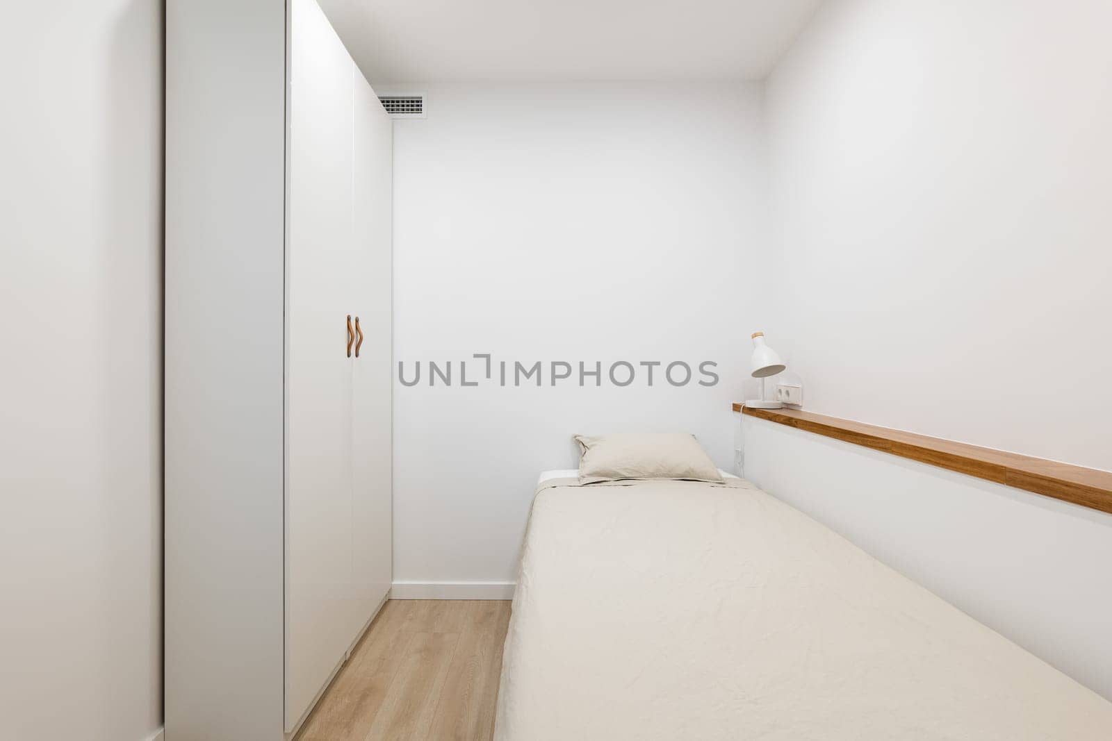 Horizontal shot of a single bed with a spacious white wardrobe, shelf and light fixture against white walls. Concept of an apartment bought with a mortgage for a single person by apavlin