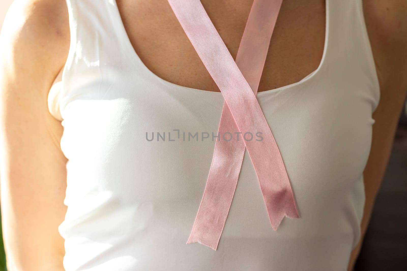 Shot of the woman in the white top against the white wall, with pink ribbon on her neck as a symbol of breast cancer awareness
