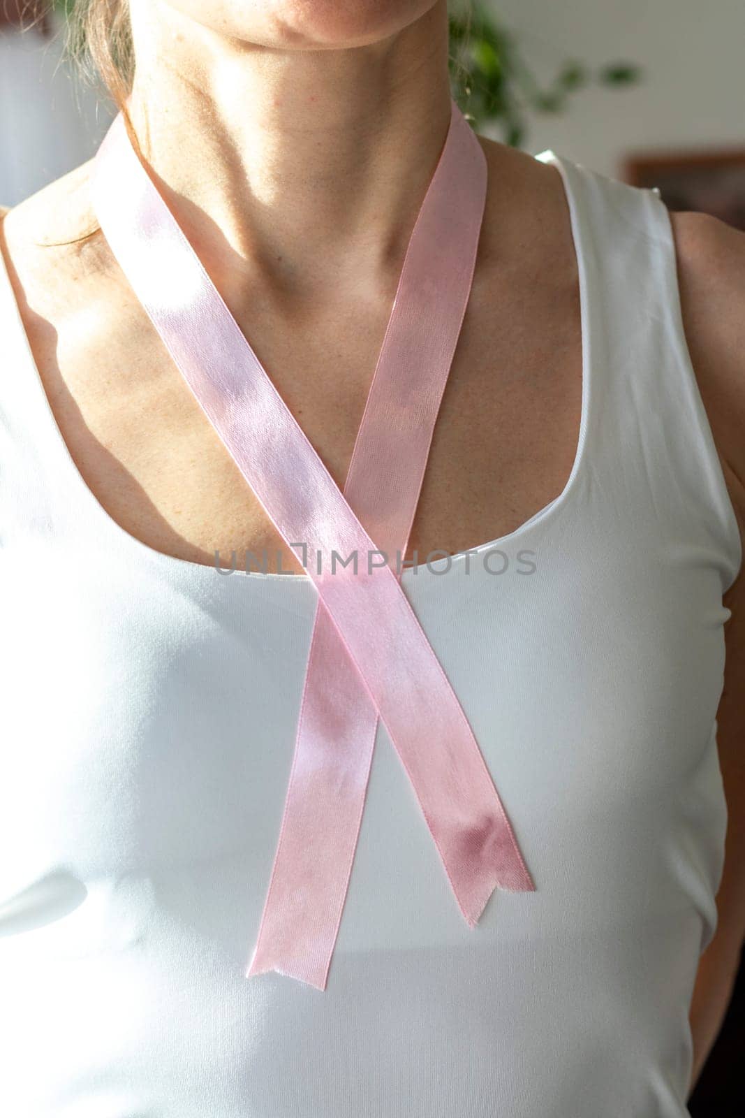 Shot of the woman in the white top against the white wall, with pink ribbon on her neck as a symbol of breast cancer awareness