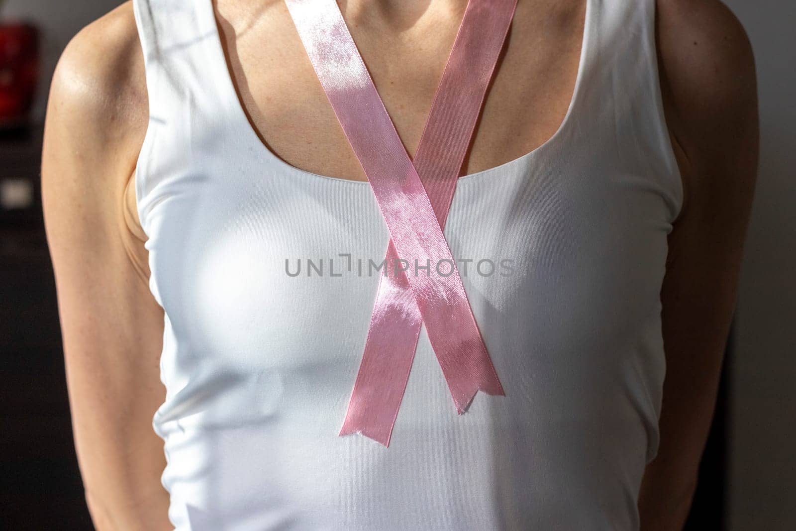 Shot of the woman in the white top against the white wall, with pink ribbon on her neck as a symbol of breast cancer awareness