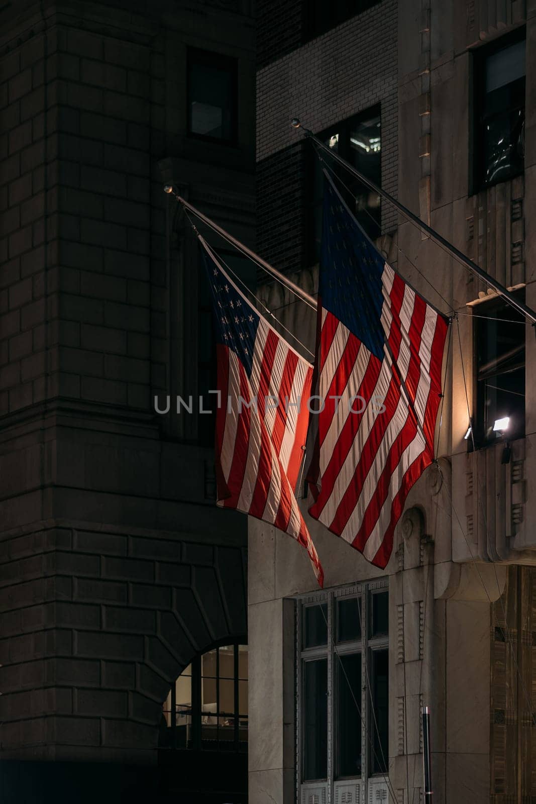 Two American flags wave in the night, anchored on an urban building.