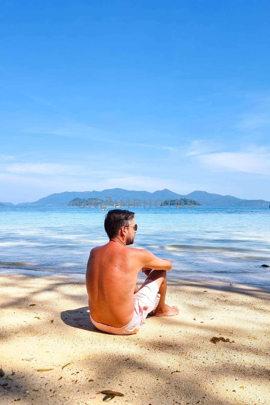 A man in casual clothing sits on the sandy beach, his gaze fixed out to the vast expanse of the ocean before him by fokkebok