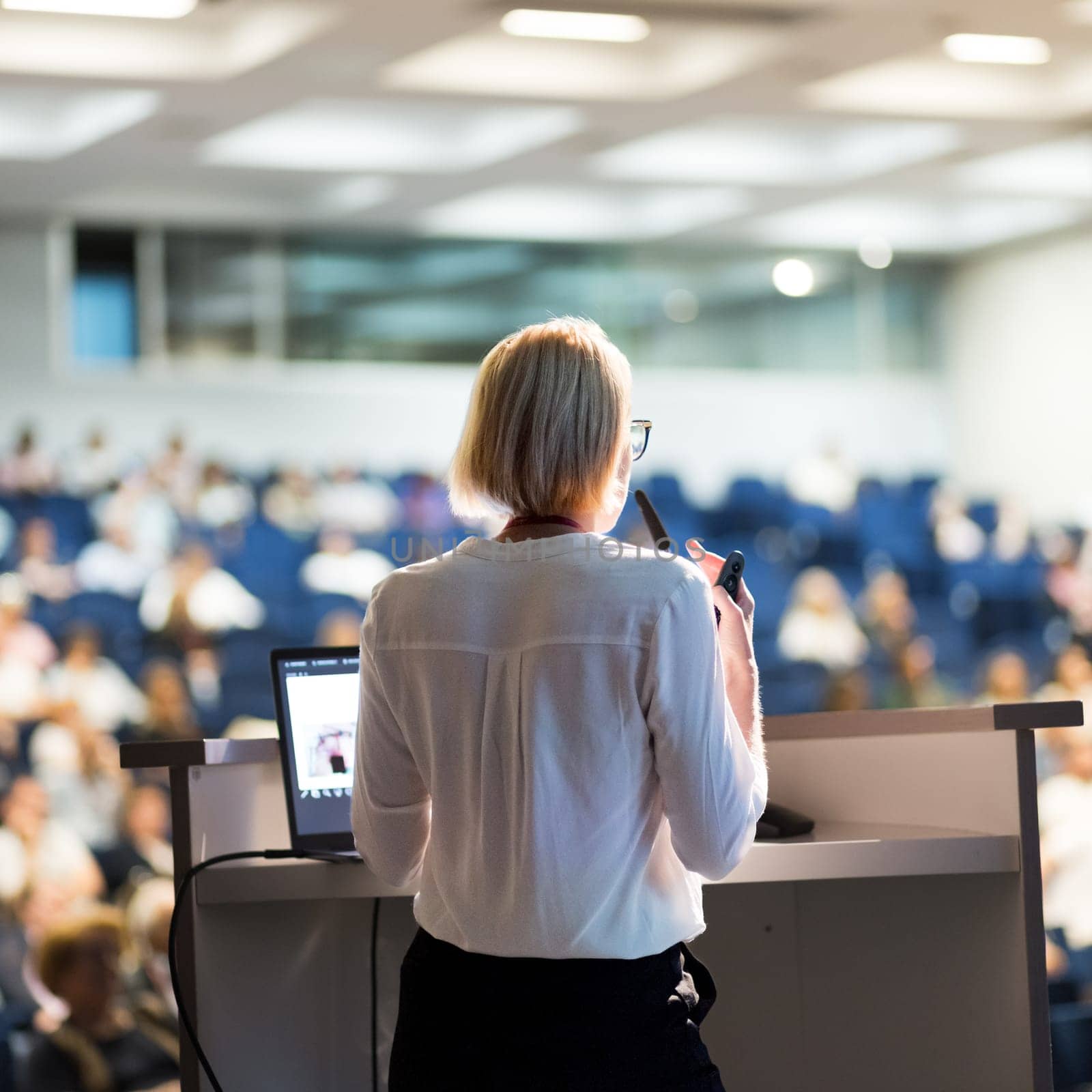 Female speaker giving a talk on corporate business conference. Unrecognizable people in audience at conference hall. Business and Entrepreneurship event