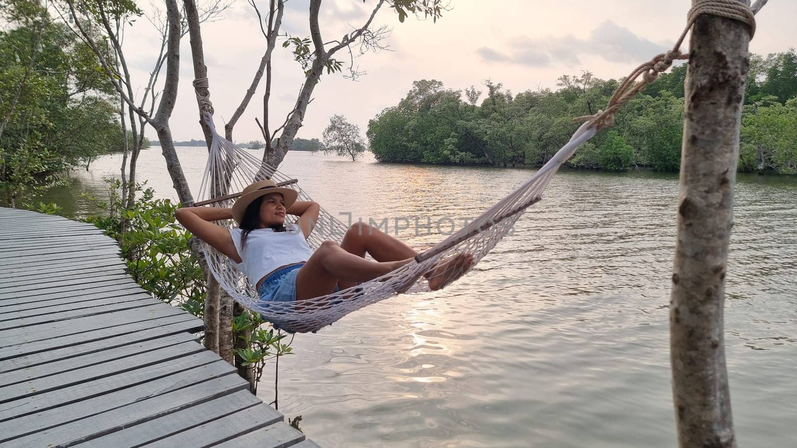 A woman peacefully relaxes in a hammock, gently swaying back and forth on a wooden dock surrounded by water. Chantaburi Thailand