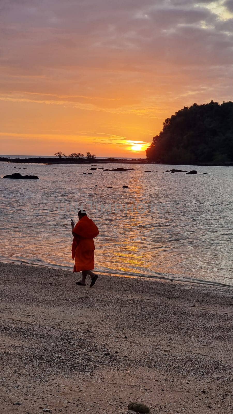 A lone Thai monk figure strolls along the sandy beach as the sun sets, creating a mesmerizing silhouette against the colorful sky. Koh Libong Thailand