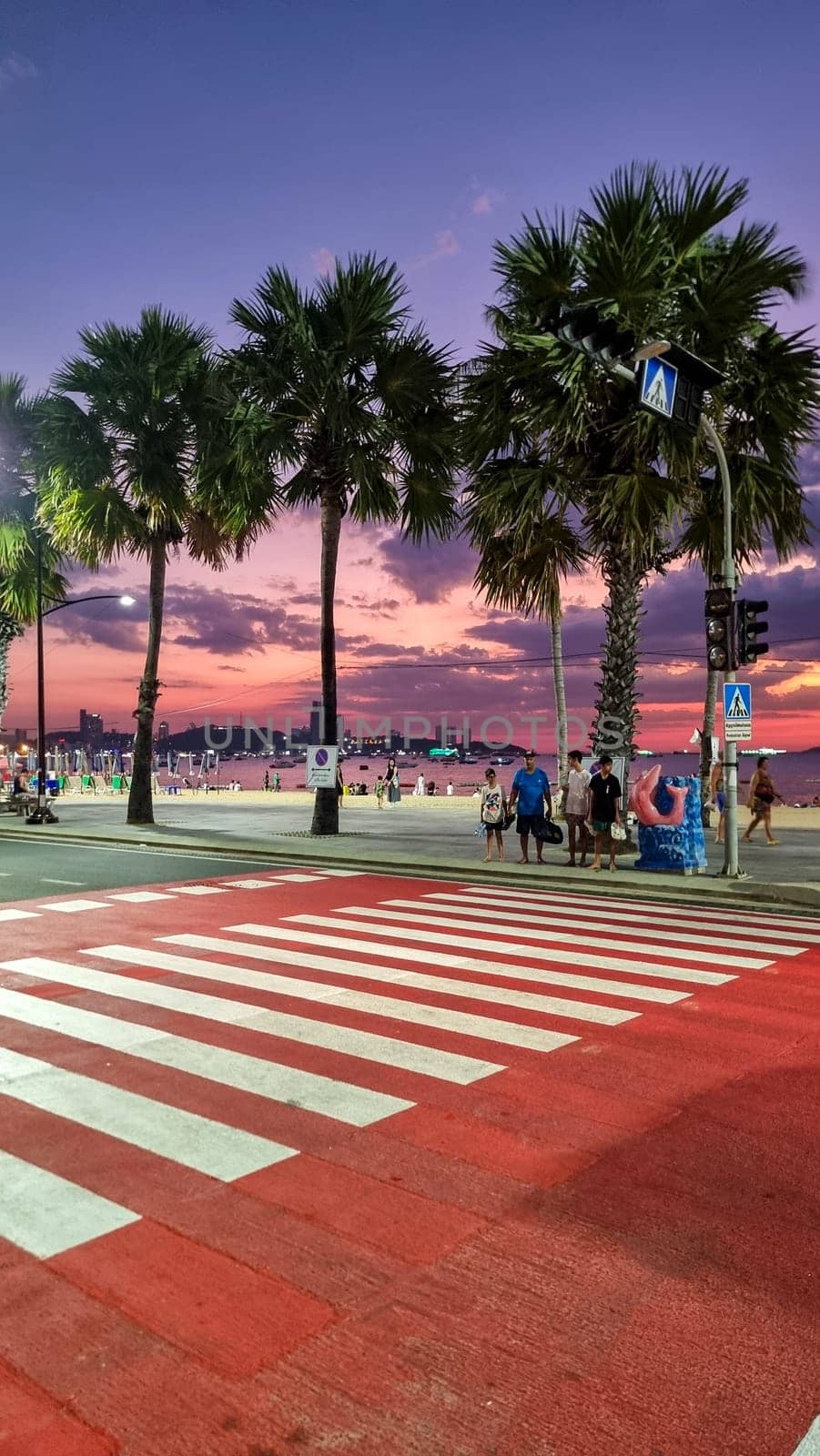 Pattaya Thailand 19 December 2024, A city street lined with towering palm trees, with a crosswalk in the foreground where pedestrians cross.