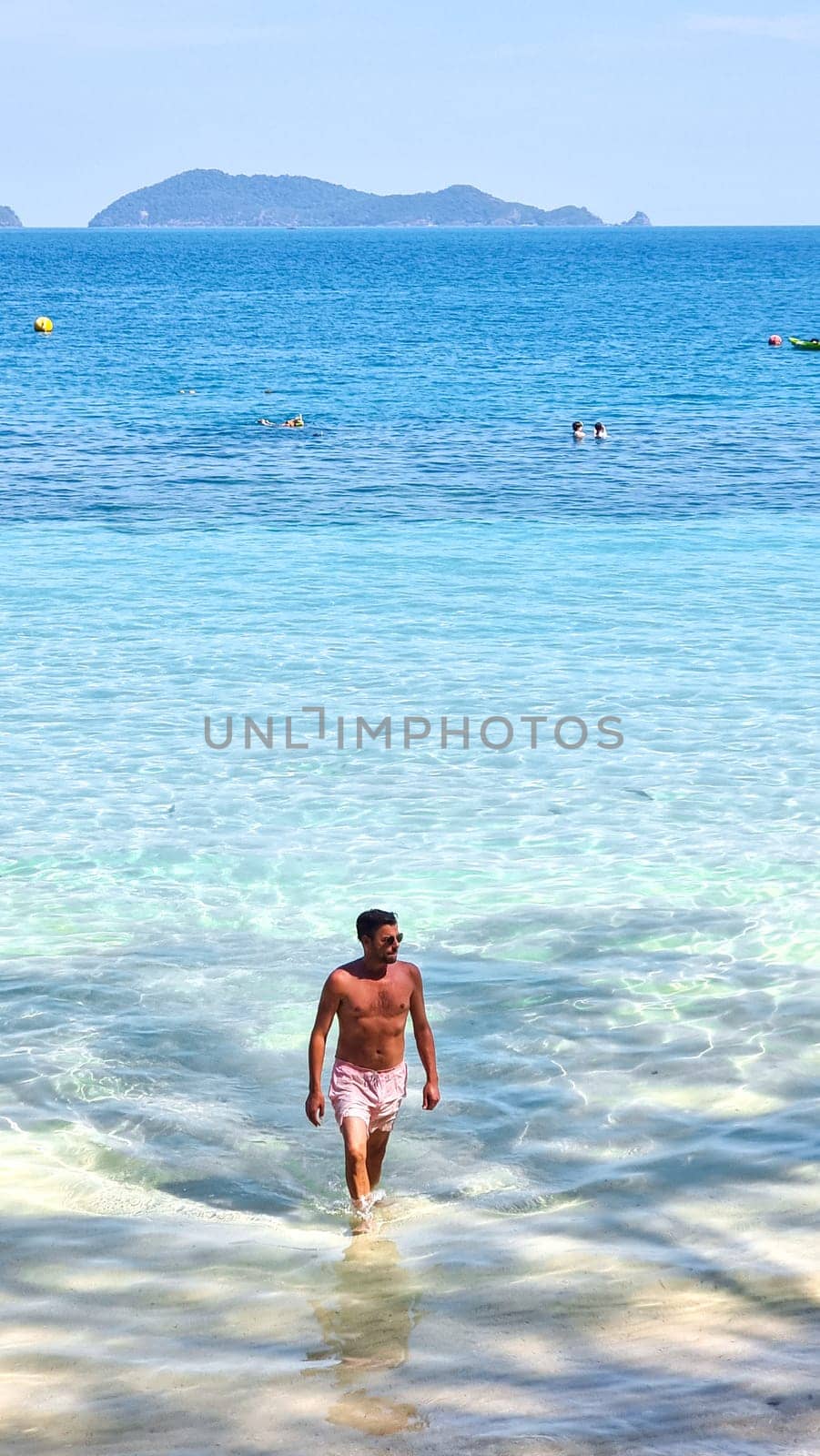 A man with a rugged appearance walks gracefully out of the glistening ocean water, the droplets sparkle in the sunlight as he emerges onto the sandy beach. Koh Wai Thailand
