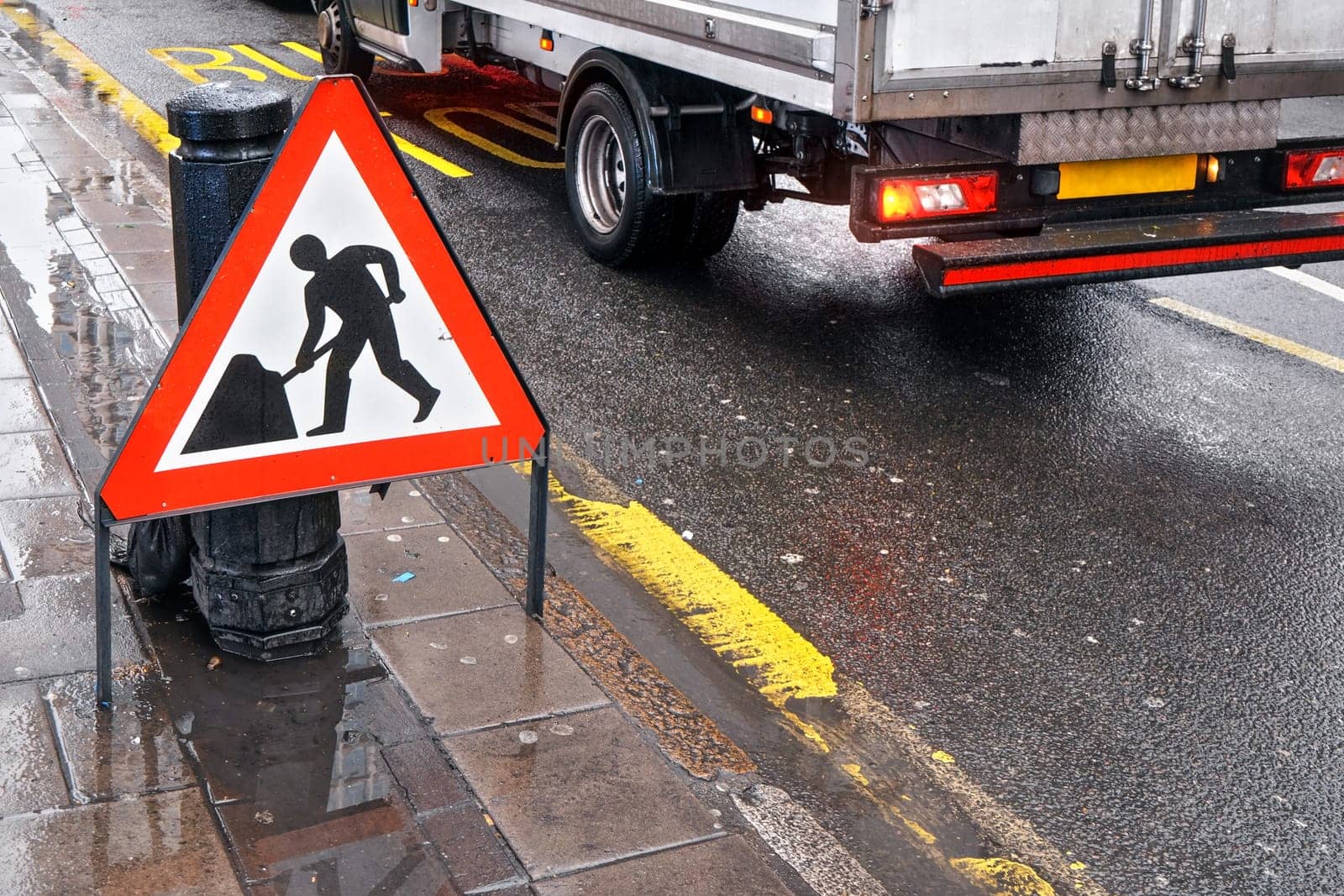 Warning roadworks sign at wet road next to pavement, van moving in background over Bus Stop writing on road by Ivanko