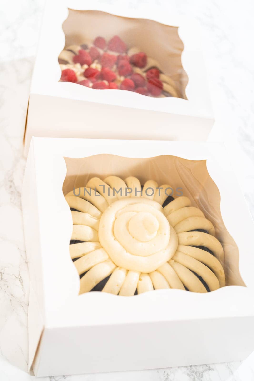 Two delightful homemade bundt cakes, each adorned with creamy cream cheese frosting, elegantly packaged in white paper boxes with a clear window to showcase their scrumptious appeal.