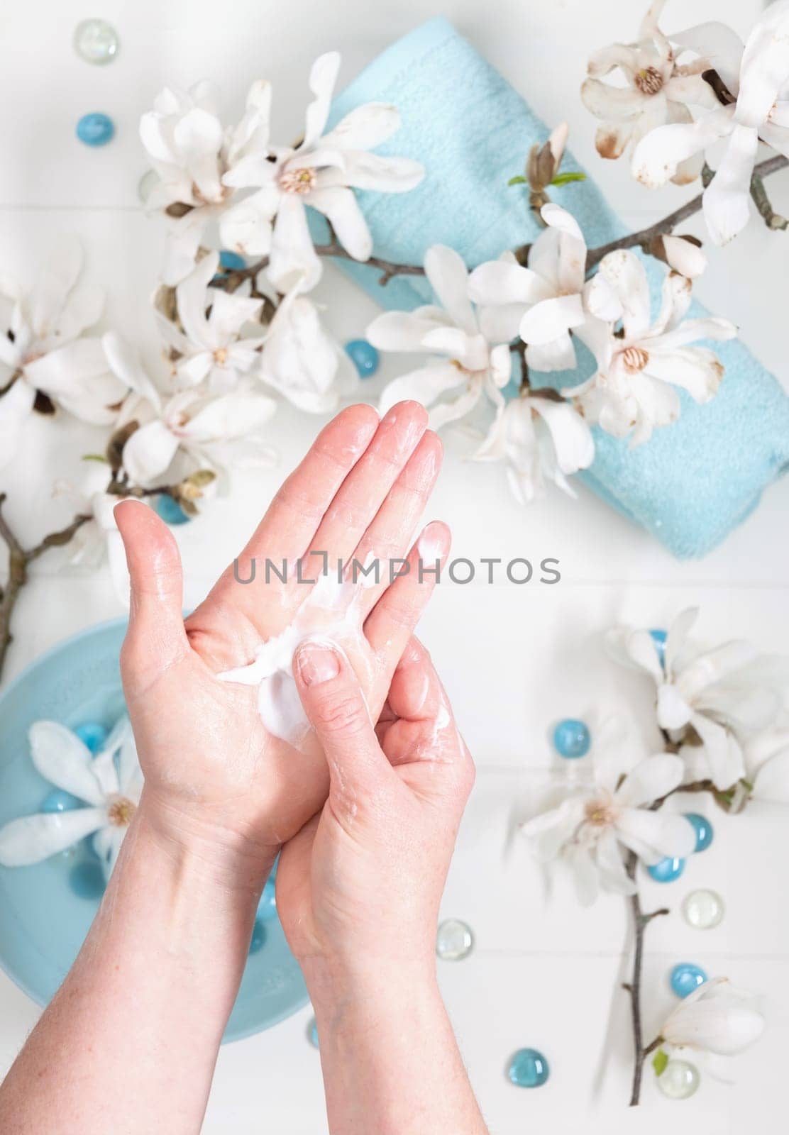 young woman doing step by step hand massage with cream in salon with cup of spring water and white magnolia flowers by KaterinaDalemans
