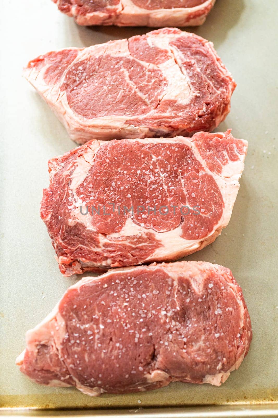 Situated in a modern white kitchen, a seasoned rib eye steak, boasting its beautiful marbling, sits ready on a baking sheet. It is prepared for the outdoor gas grill, promising a perfect sear on the exterior and a juicy, tender interior, heralding an upcoming indulgent feast.