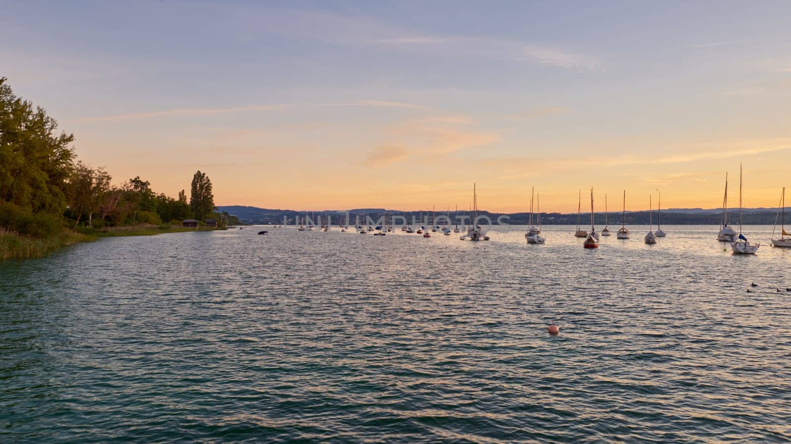 Bodensee Lake Panorama. Evening, twilight, setting sun, picturesque landscape, serene waters, boats and yachts at the dock, beautiful sky with clouds reflecting in the water, riverside at dusk, showcasing the coexistence of technology and production with the environment.