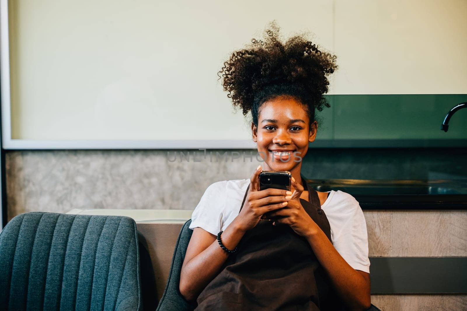 A cheerful black woman barista and coffee shop owner in uniform using a mobile phone. Portrait of the smiling entrepreneur working and communicating joyfully. by Sorapop