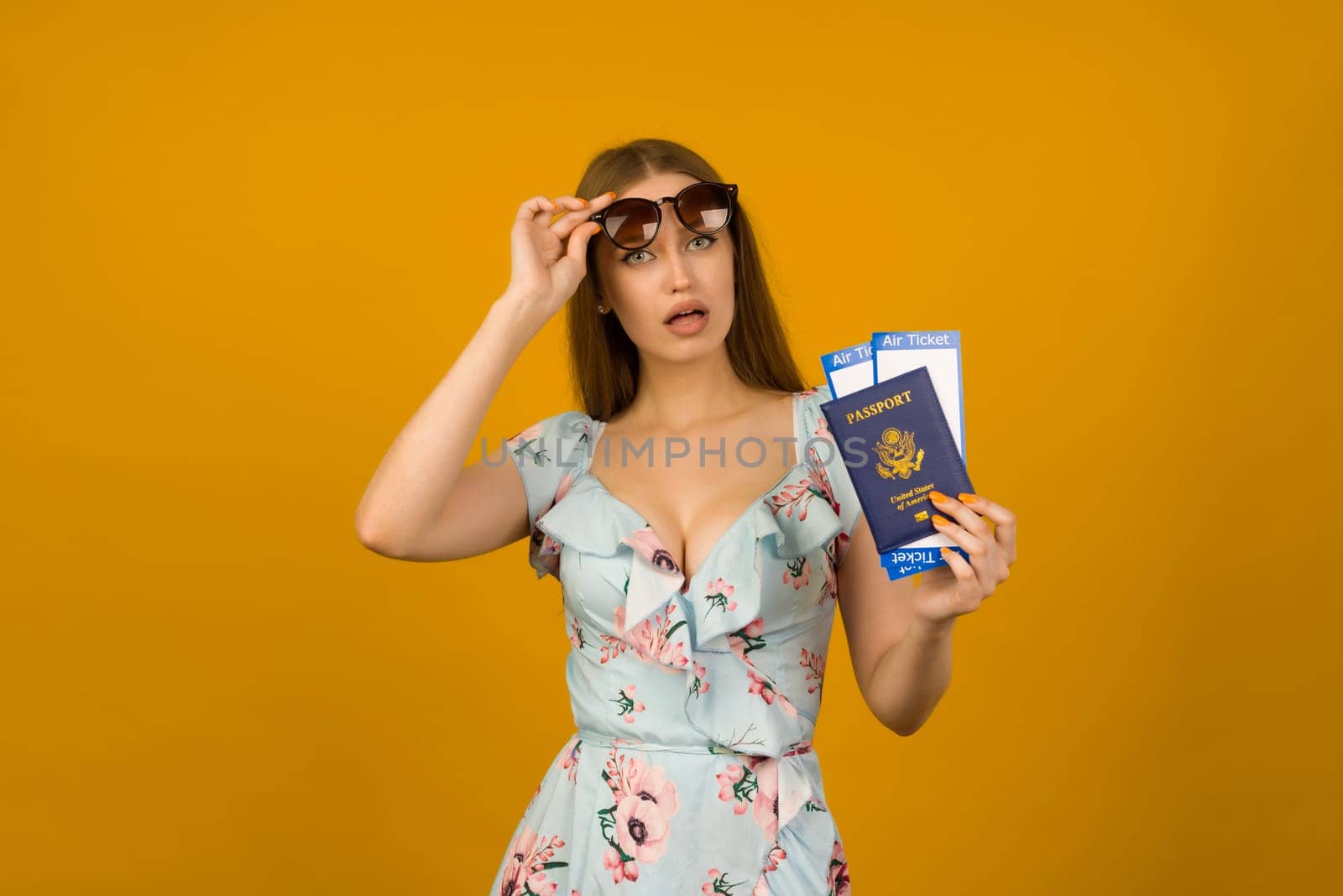 Pop-eyed young woman in blue dress with flowers and sunglasses is holding airline tickets with a passport on a yellow background. Rejoices in the resumption of tourism after the coronovirus pandemic.