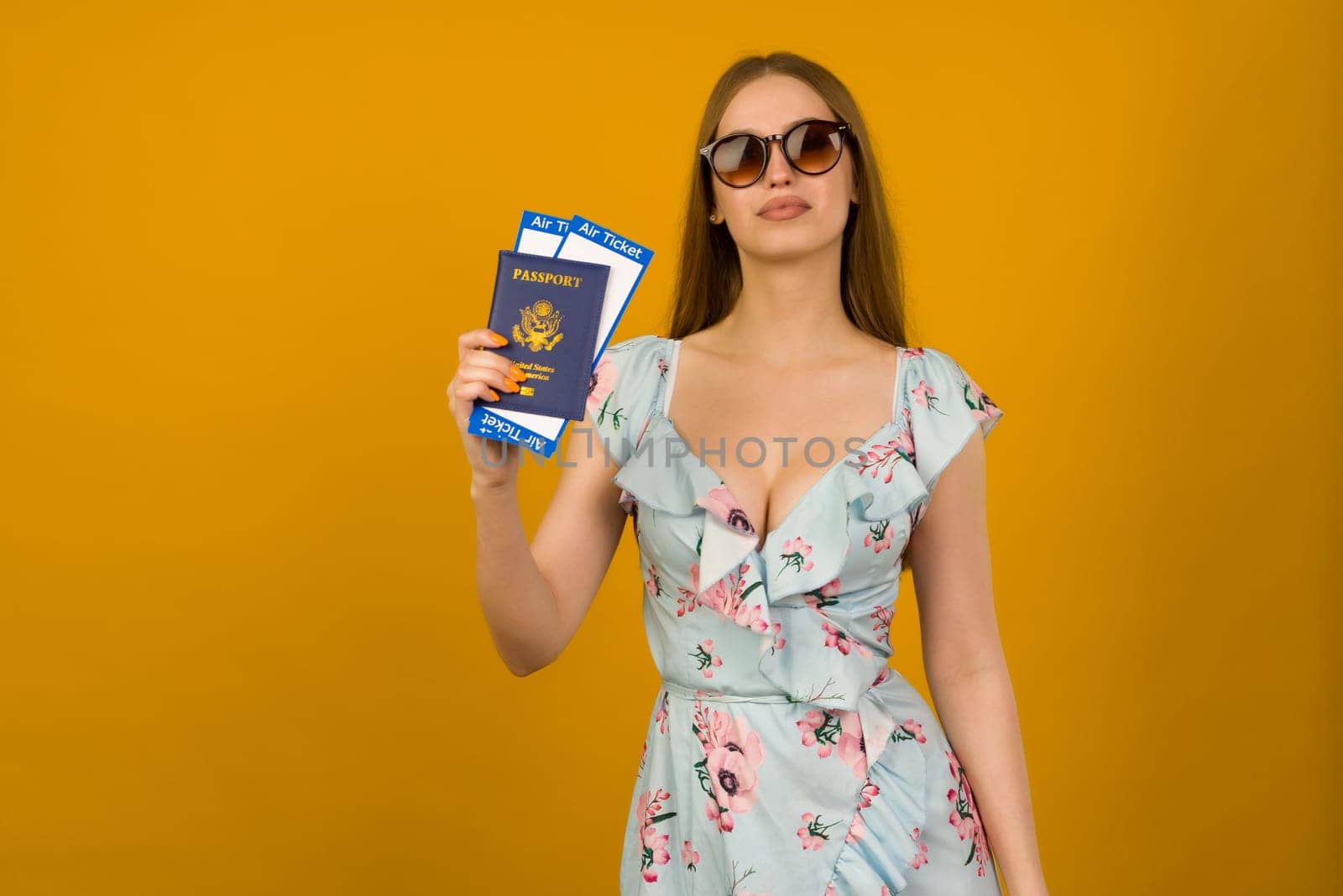 Joyful young woman in blue dress with flowers and sunglasses is holding airline tickets with a passport on a yellow background. by zartarn