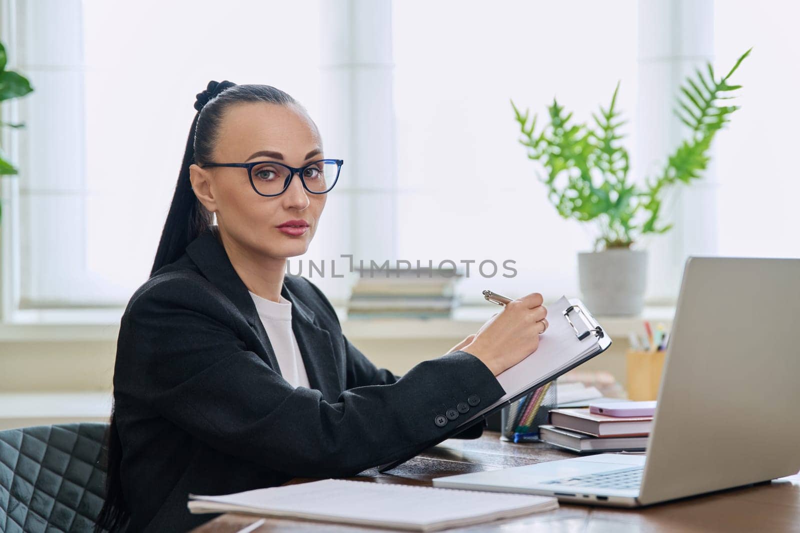 Portrait of businesswoman with clipboard sitting at workplace with computer laptop. Confident 30s female looking at camera. Remote business, technologies internet, teaching, blogging, work freelance