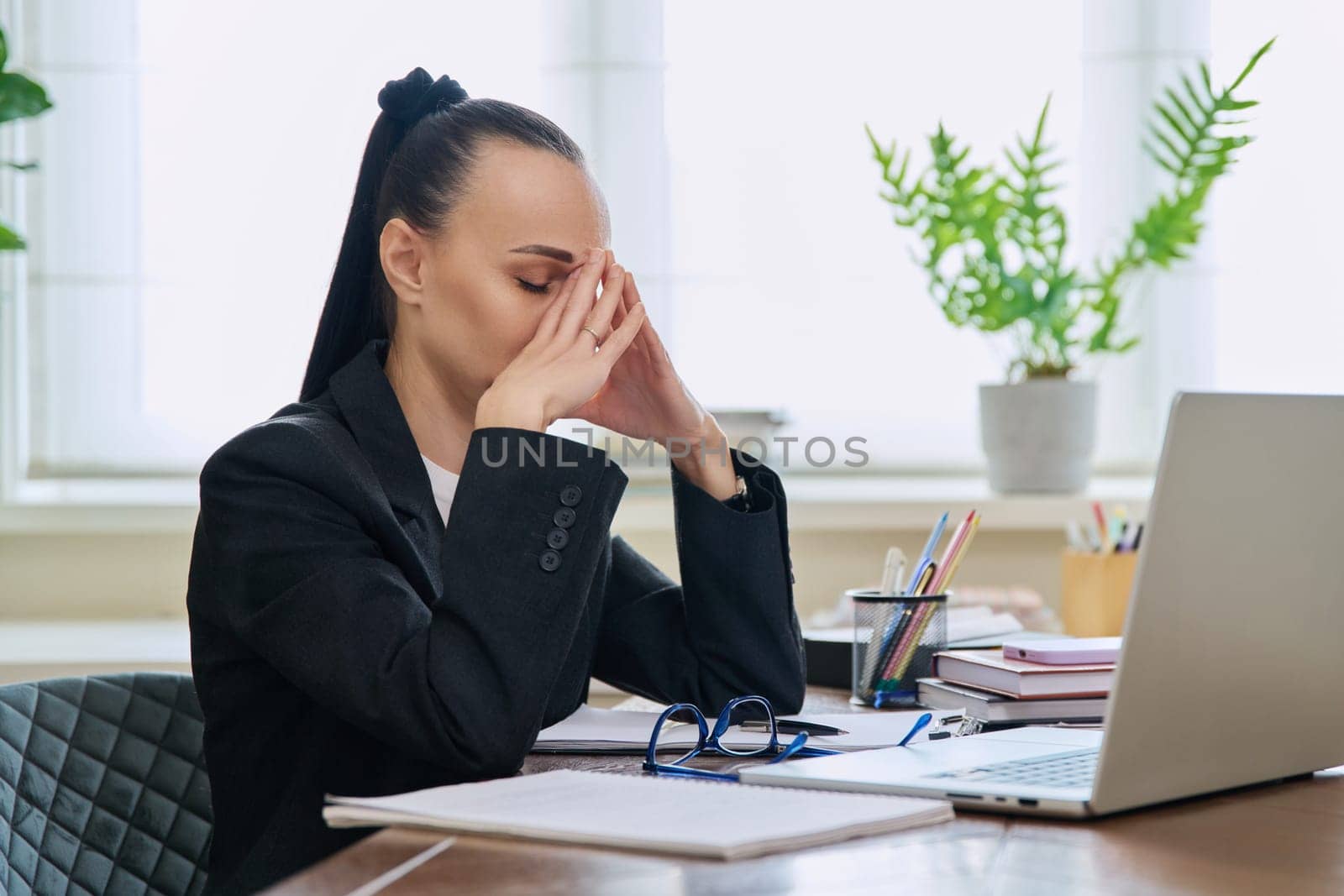 Tired sad business woman at the workplace. Upset female sitting at desk with laptop computer, experiencing stress pain headache. Business, remote work, health problems, freelance concept