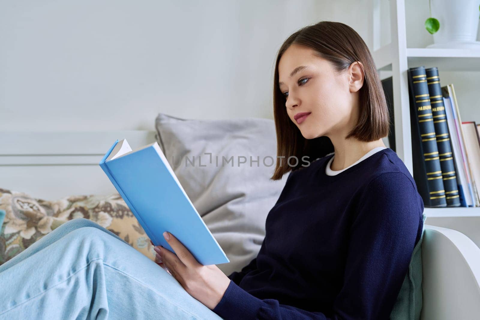 Young woman reading paper book, relaxed sitting on sofa at home. Fiction, hobby, leisure, youth concept