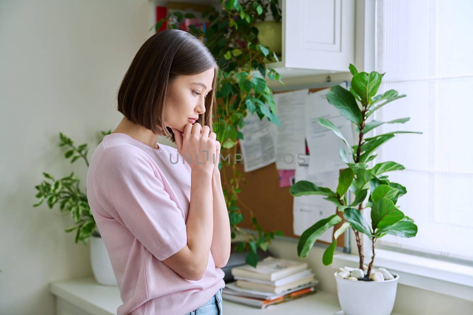 Young sad upset unhappy woman standing looking out the window at home. Frustrated confused female experiencing difficulties. Mental problems, feelings loneliness stress depression difficulties youth