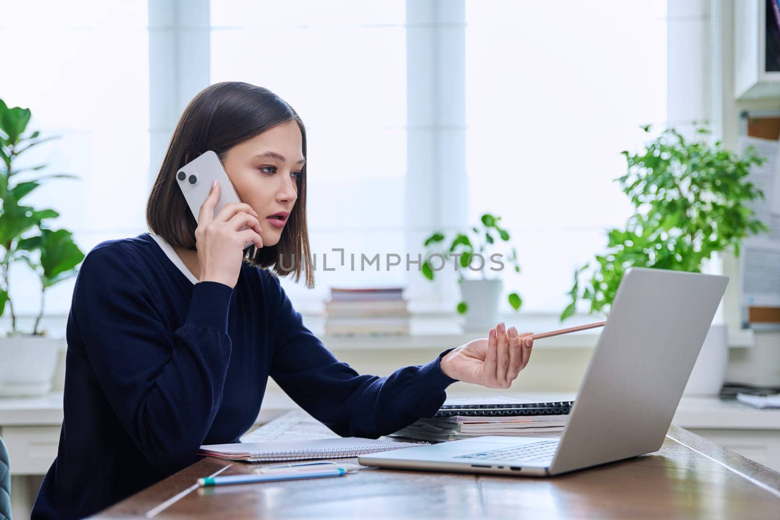 Young woman talking on mobile phone sitting at desk with laptop computer. Serious confident 20s female working, learning. Work, business, workplace, study, education, training, people concept