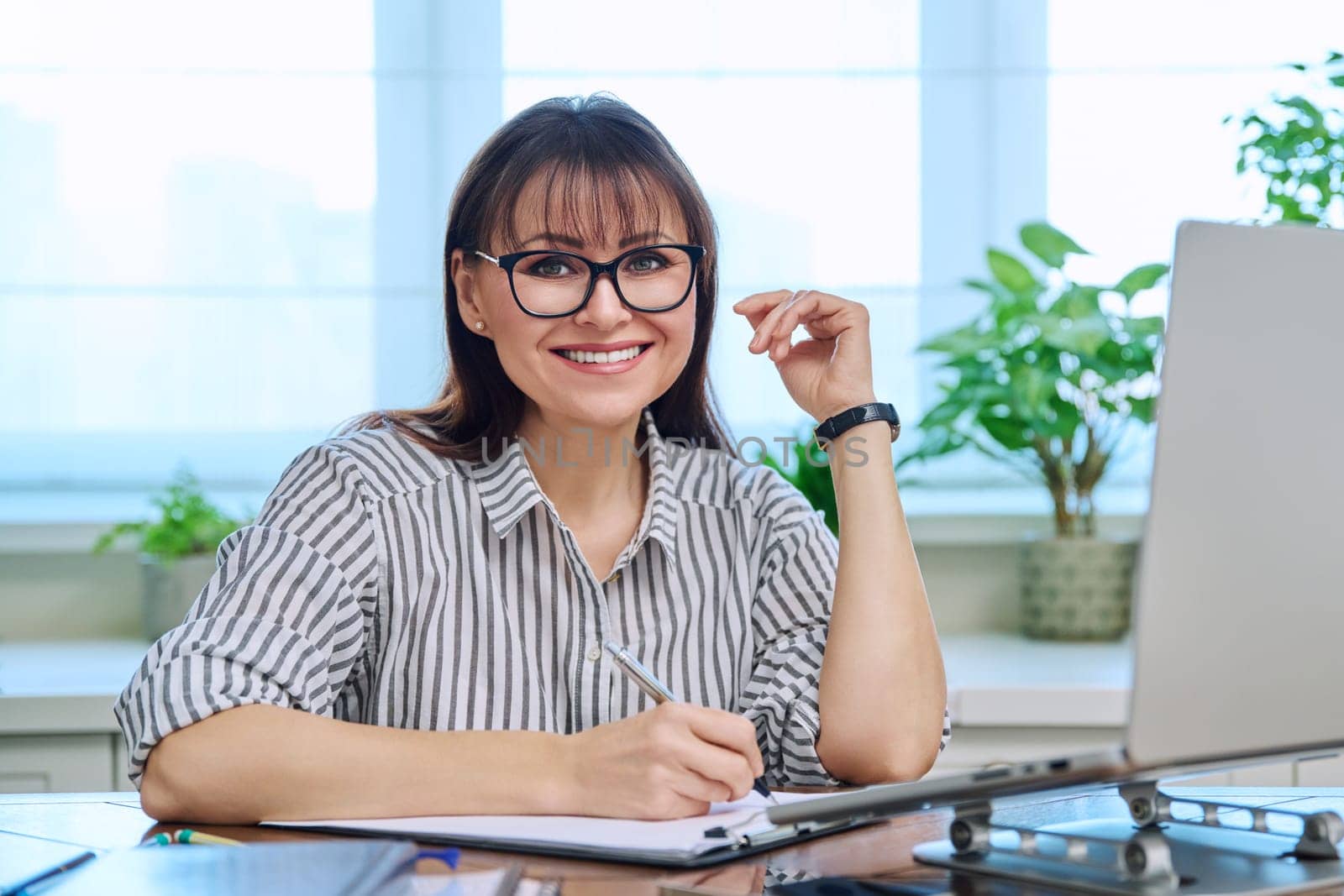 Portrait of middle-aged woman at her workplace at desk with computer, looking at camera. Mature smiling female working in home office. Remote business, teaching, work, freelancing