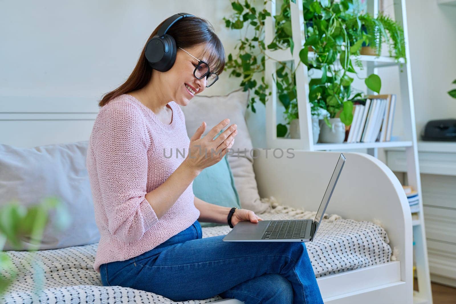 Middle aged woman in headphones having video call chat conference on laptop sitting on couch at home. Vlog, work, leisure, online communication, modern technologies concept